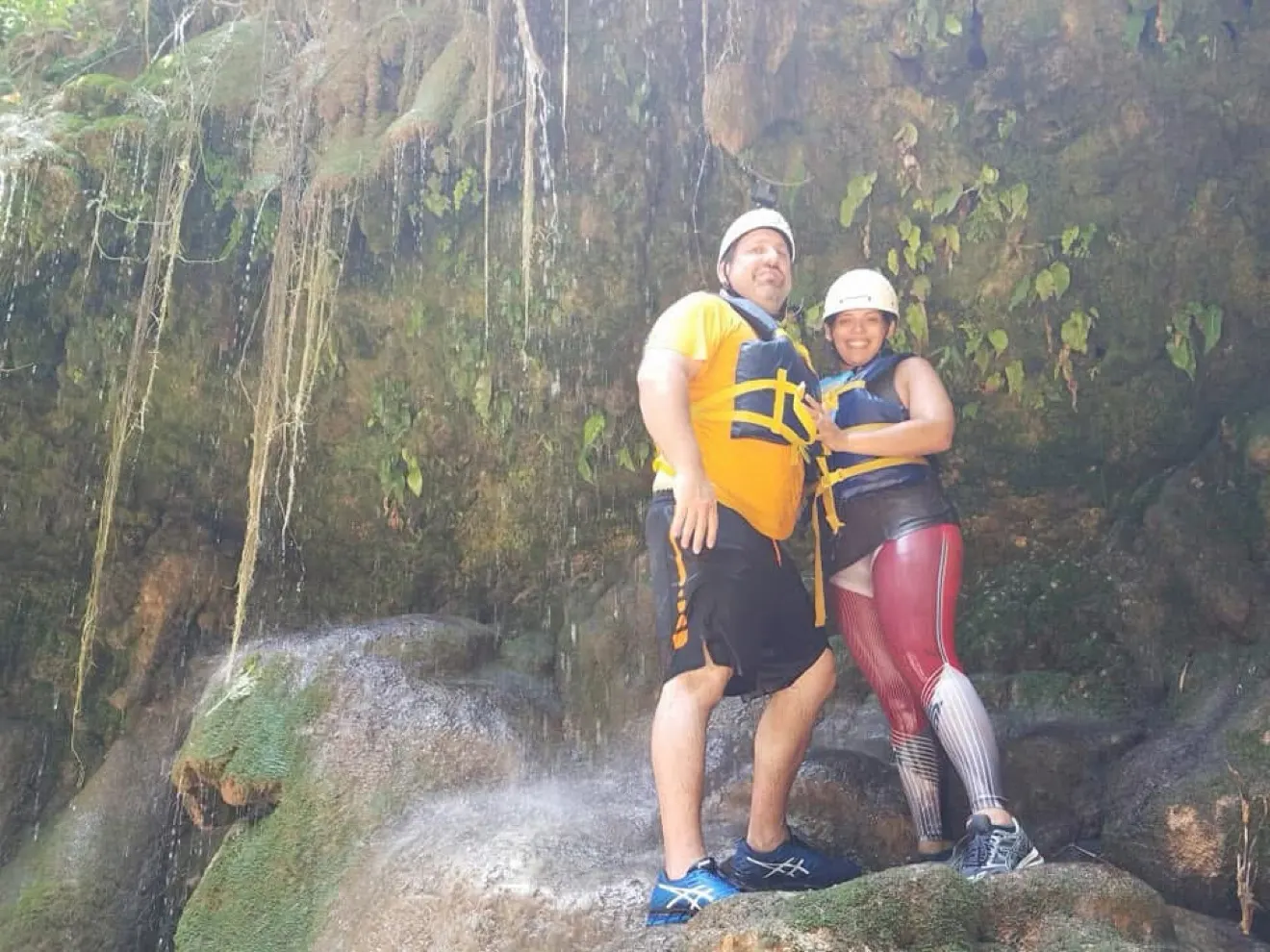 Two visitors in helmets and life vests beside a small waterfall