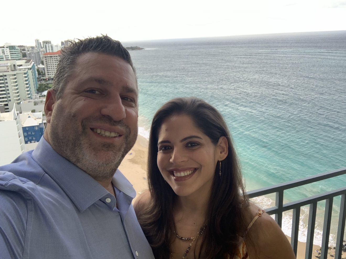 Mike and Neri smiling together on a balcony overlooking Isla Verde beach